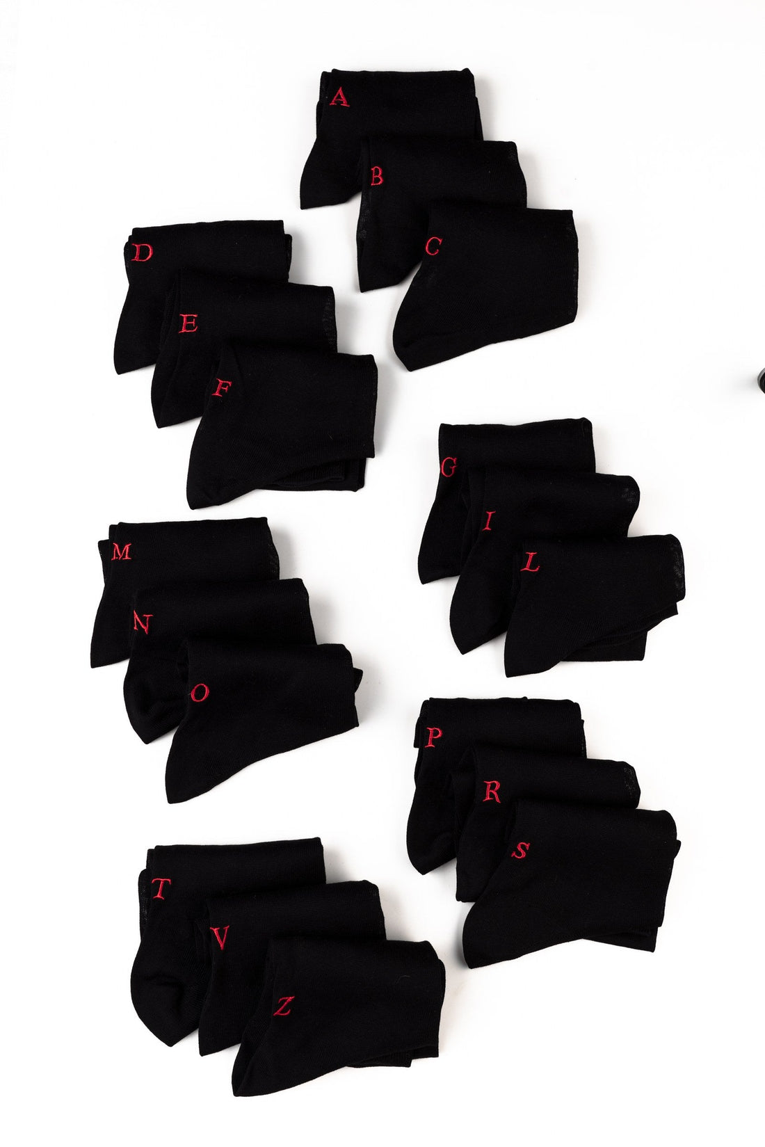 regali di Natale per i colleghi: calze nere personalizzate con iniziali diverse ricamate in rosso, disposte a gruppi su fondo bianco