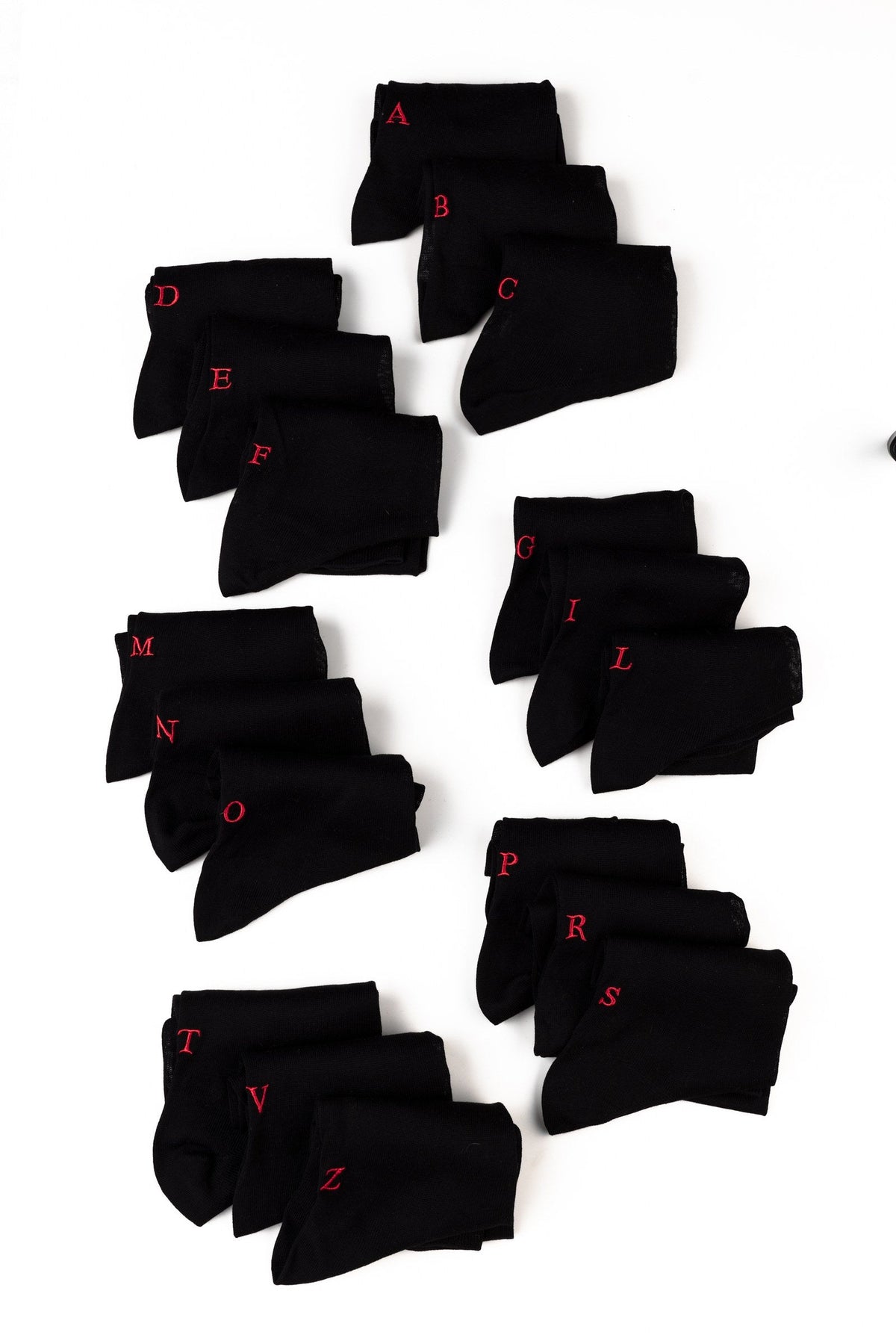 regali di Natale per i colleghi: calze nere personalizzate con iniziali diverse ricamate in rosso, disposte a gruppi su fondo bianco